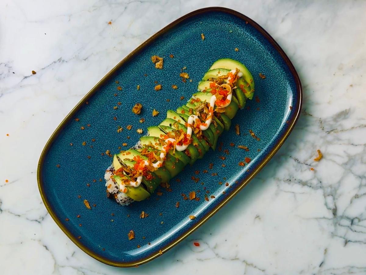 A serving platter with a full roll of vegan caterpillar sushi, topped with avocado strips, vegan kewpie mayo, vegan caviar and dried onion flakes.