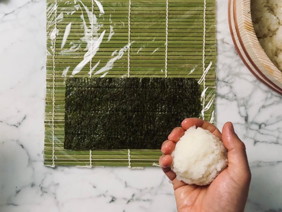 A wet hand holding a ball of sushi rice, roughly the size of a medium apple, positioned above the prepared nori sheet on the sushi mat.
