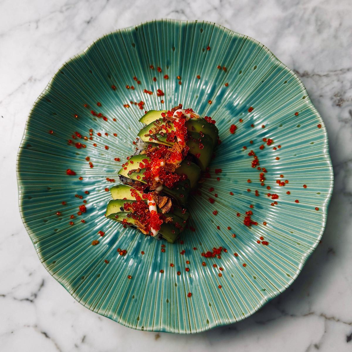 Vegan sushi roll presented on a serving dish, topped with avocado, vegan caviar, vegan kewpie mayo and dried onion flakes.