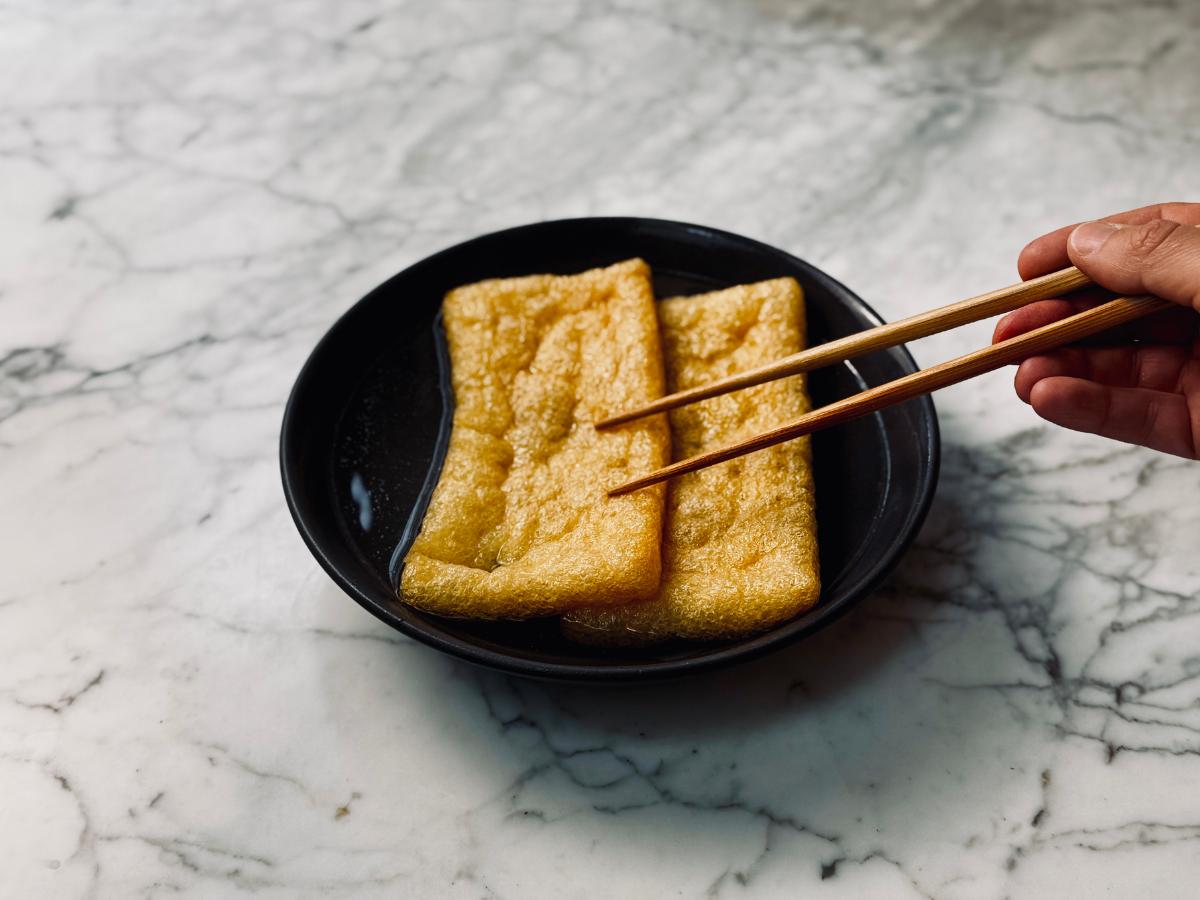 Fried tofu sheets rest in a bowl of shallow liquid, with some chopsticks gently dunking them under.