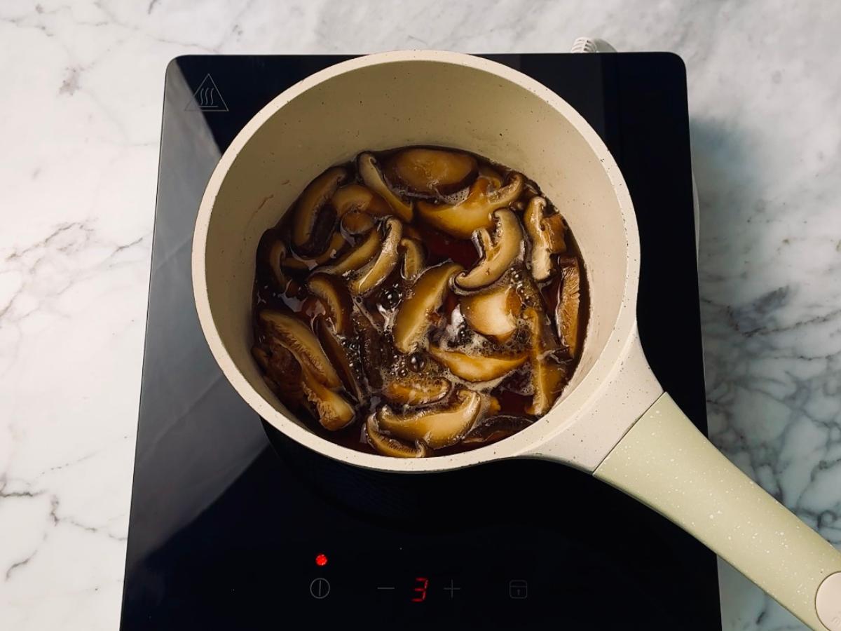 A small saucepan of sliced shiitake mushroom strips in liquid, on stovetop heat.