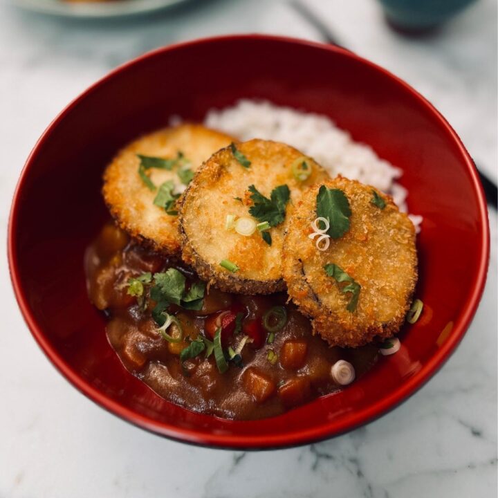 A close-up shot of a bowl of katsu curry, showing the contrast between crunchy breaded aubergine cutlets and rich, glossy curry sauce over fluffy rice, with fresh garnishes adding color.