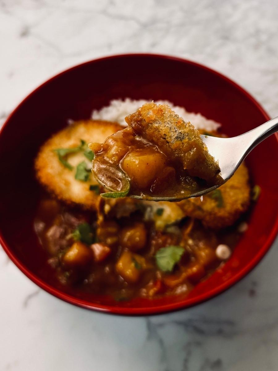 A spoonful of curry and aubergine katsu held up close to the camera, capturing the texture of the crispy coating and saucy curry in one bite with the bowl of katsu curry and rice in the background.