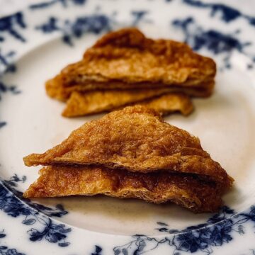 Close-up of two golden-brown inari age tofu pouches, folded into neat triangles and arranged on a white plate with a blue floral rim. The surface is slightly glossy, highlighting their layered, fried texture.