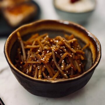 Small ceramic bowl filled with kinpira gobo - shredded burdock root and carrot sautéed in soy sauce and mirin - topped with a sprinkle of toasted sesame seeds.