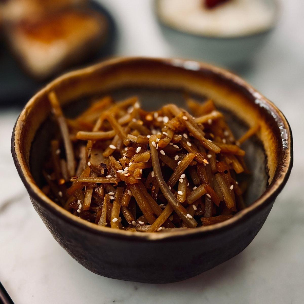 Small ceramic bowl filled with kinpira gobo - shredded burdock root and carrot sautéed in soy sauce and mirin - topped with a sprinkle of toasted sesame seeds.