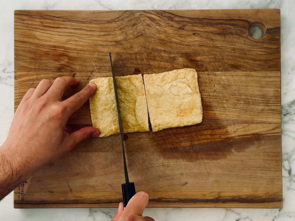 Abura-age being sliced into four sections on a wooden cutting board.
