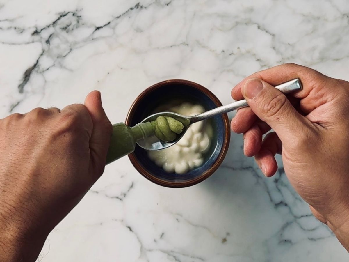 A pair of hands squeezing green wasabi paste onto a teaspoon over a small bowl of mayo.