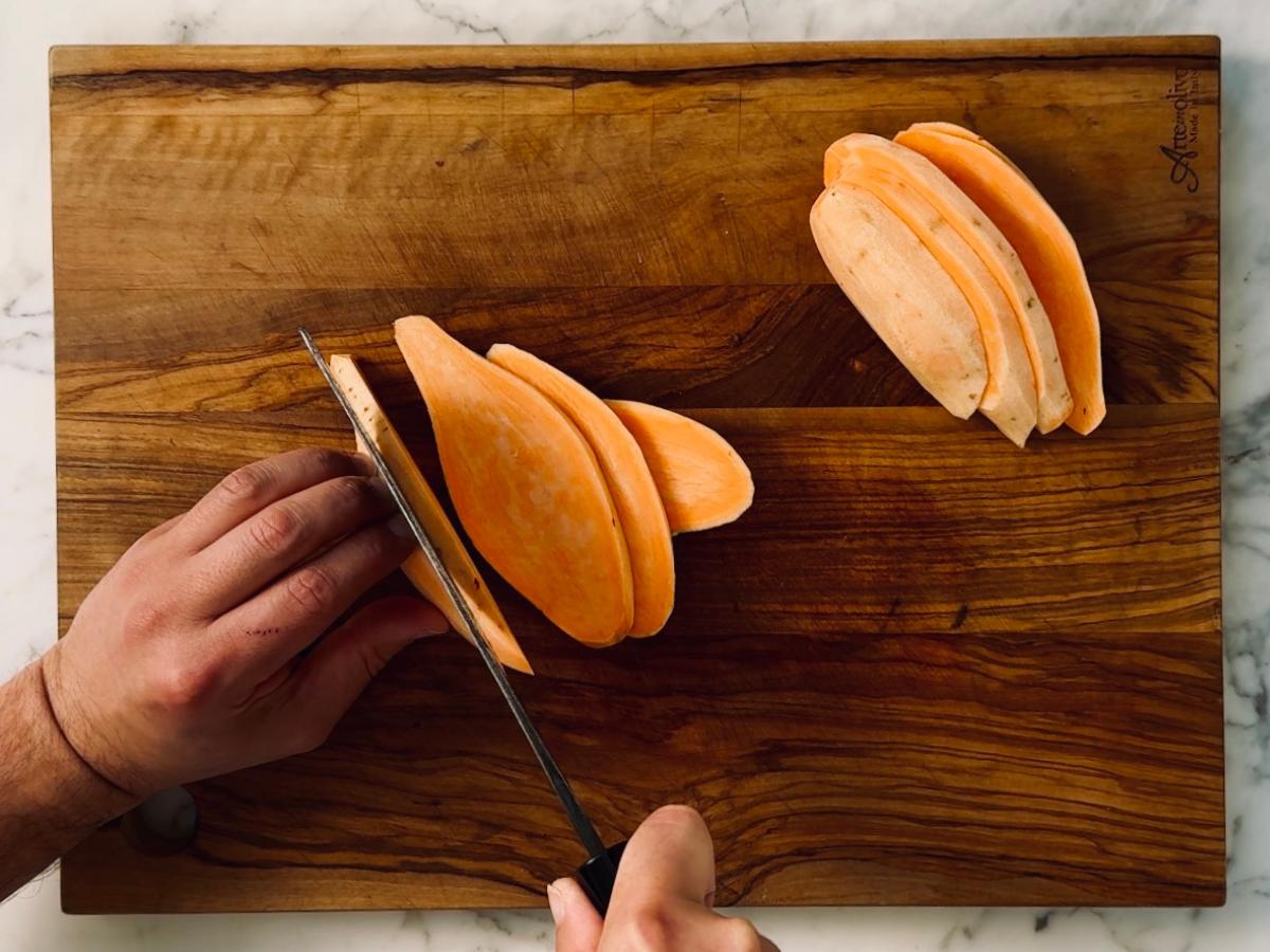 Sweet potato being sliced into ovals on a wooden cutting board, using a large sharp knife.