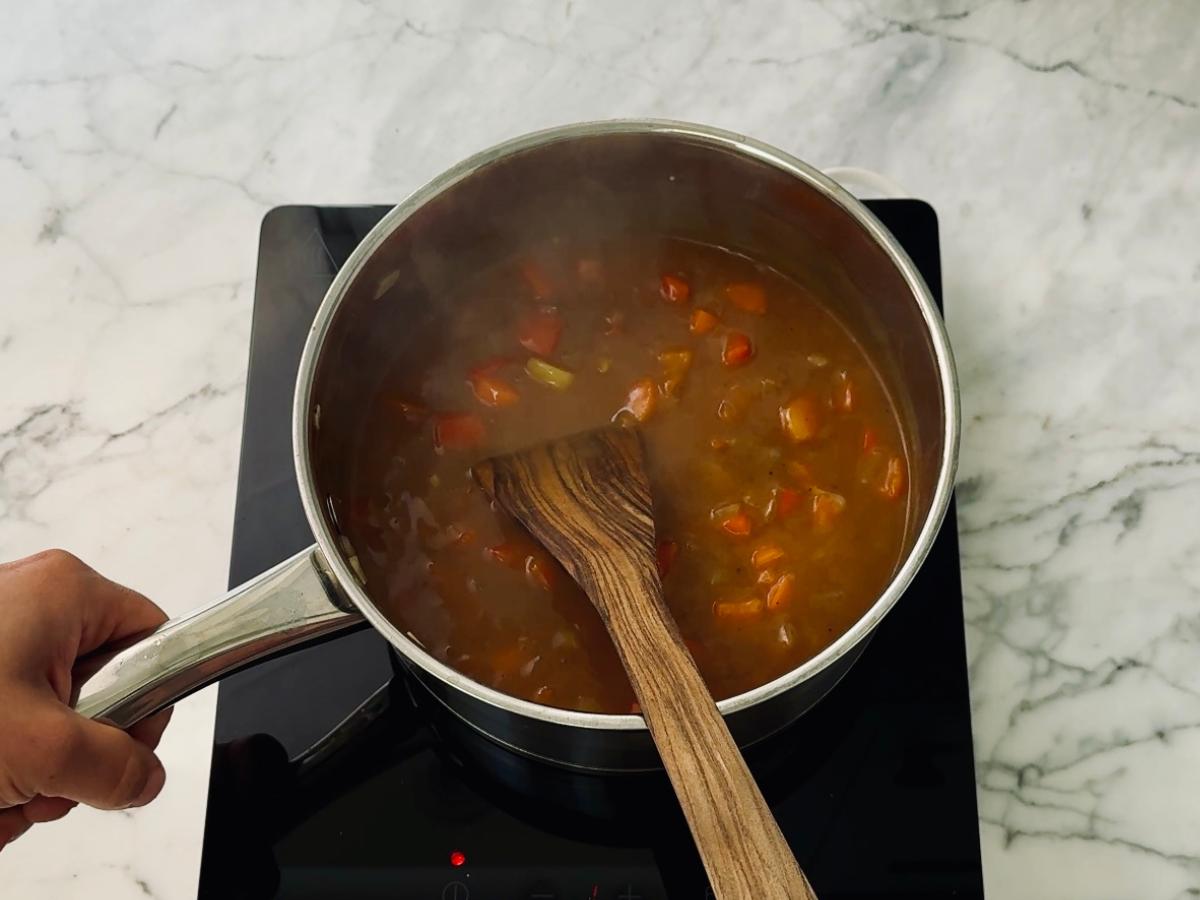 A saucepan over heat containing a dark brown sauce (from curry roux cubes) and vegetables, being stirred with a wooden paddle as it thickens to make a rich curry sauce for the katsu curry.