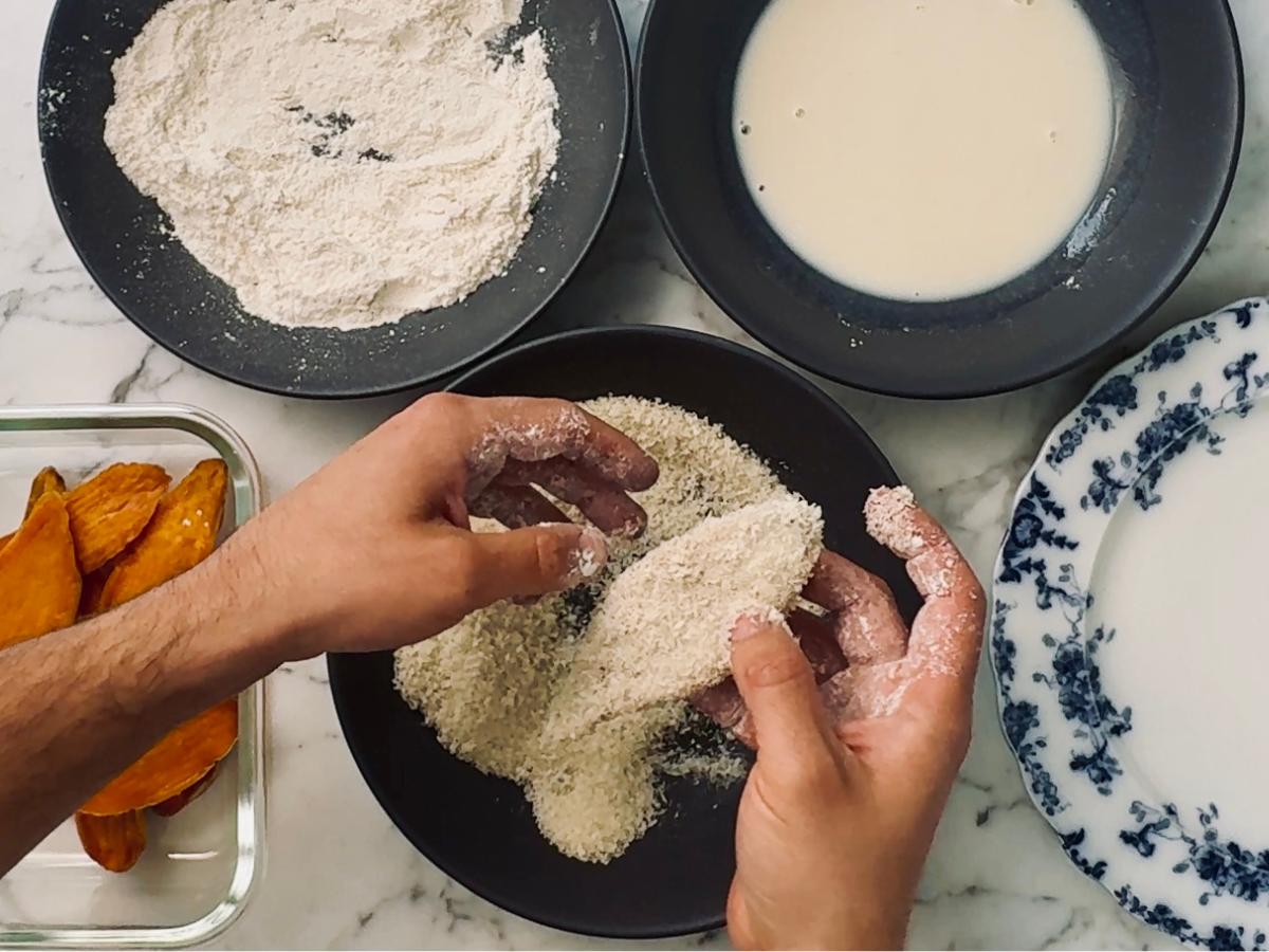 A pair of hands coating a sweet potato slice in panko breadcrumbs, ensuring it's evenly covered before placing flat, ready to fry.