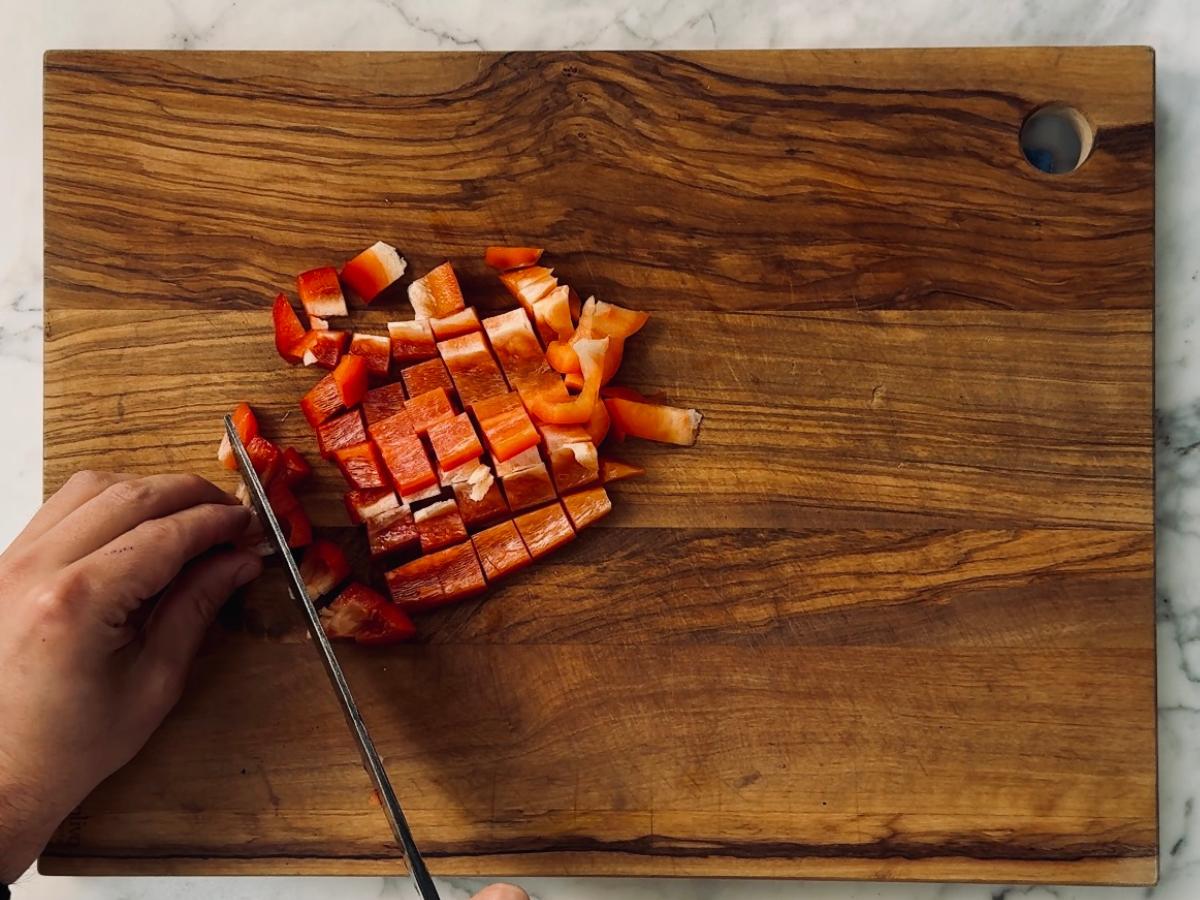 Red pepper being cut into bite-sized square pieces, using a sharp knife, on a wooden chopping board.