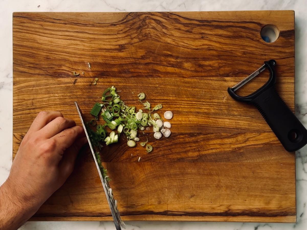 Spring onions being finely sliced on a wooden chopping board.