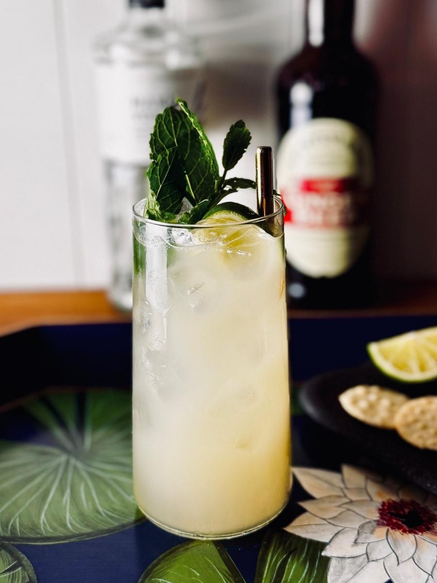 Long cocktail glass filled with Yuzu Mule, with a gradient yellow to to pale yellow colour from bottom to top; at the top of the glass floats ice, a golden steel straw, and a fresh lime and mint garnish.
