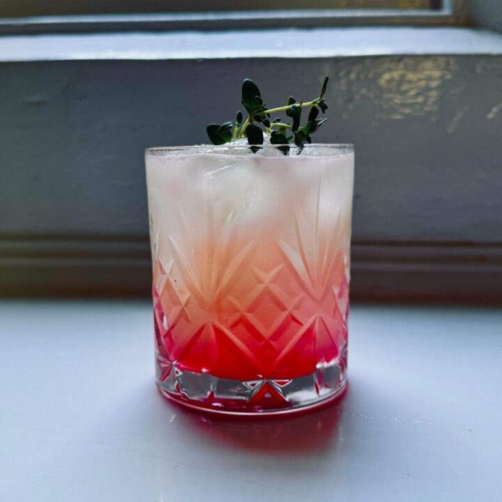 Yuzu Gin Fizz served in an old-fashioned glass over ice, showing a layered rose-to-citrus hue topped with a fresh herb garnish.