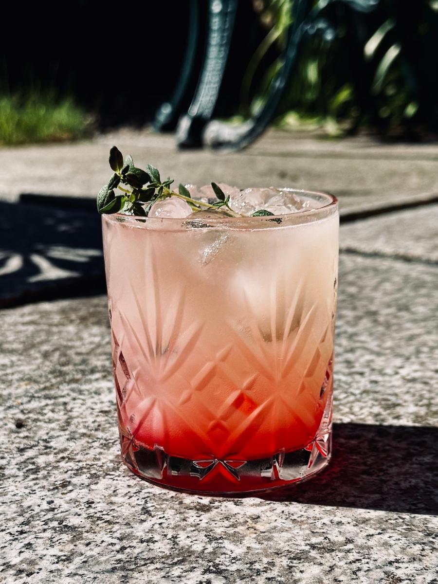 Yuzu Gin Fizz served in an old-fashioned glass over ice, showing a layered rose-to-citrus hue topped with a fresh herb garnish, with an outdoor backdrop.