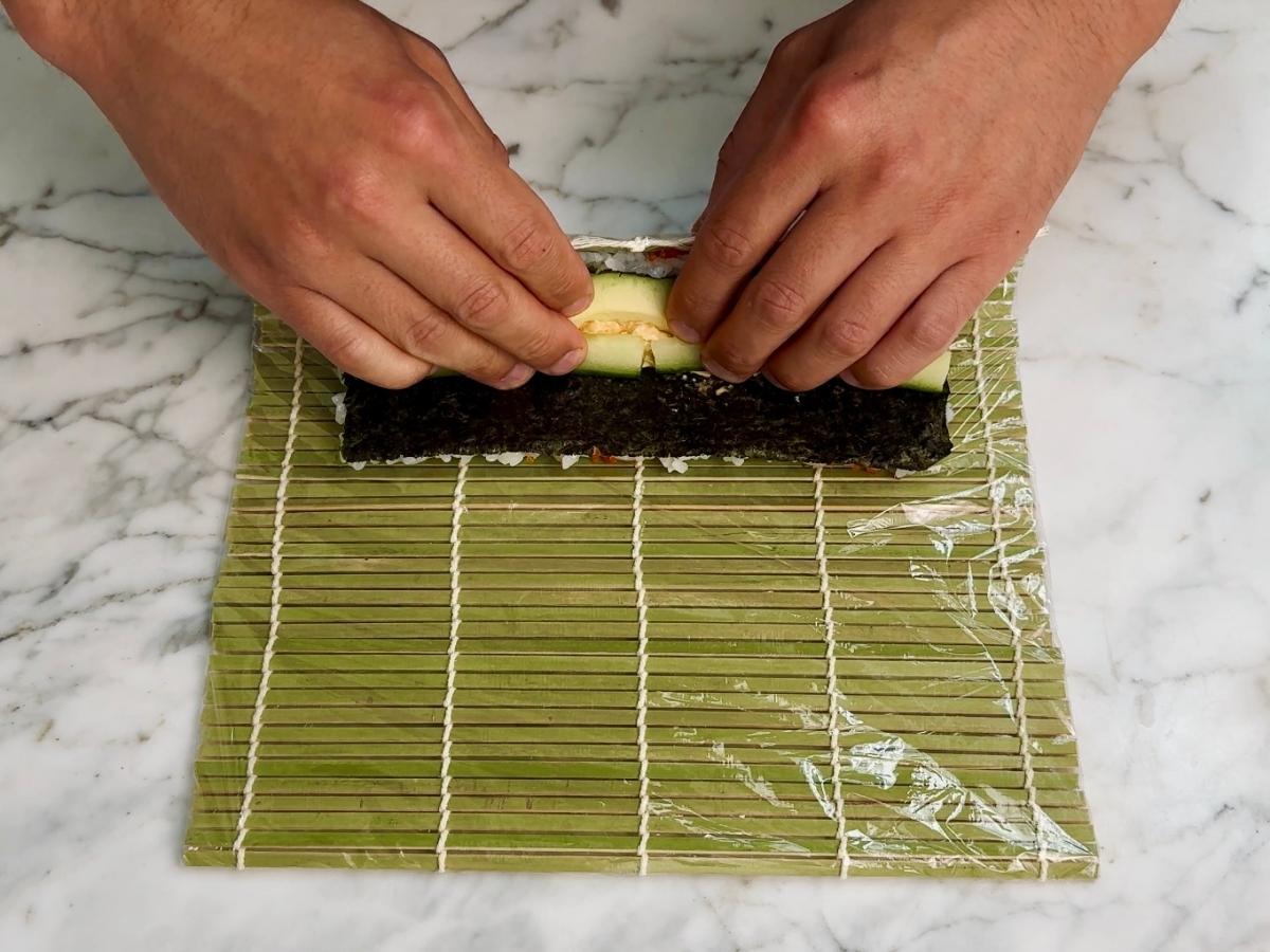 Hands folding the bamboo mat over the filling, starting the sushi roll.