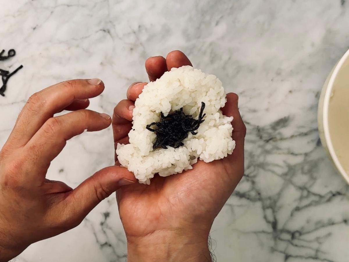 a hand holding an open rice ball with shio kombu placed centrally, on top.