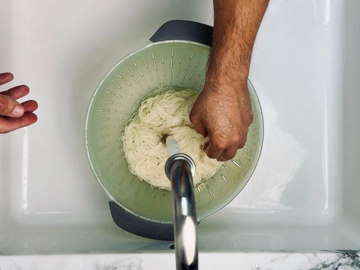 Cooked noodles being rinsed under cold running tap water in a strainer.