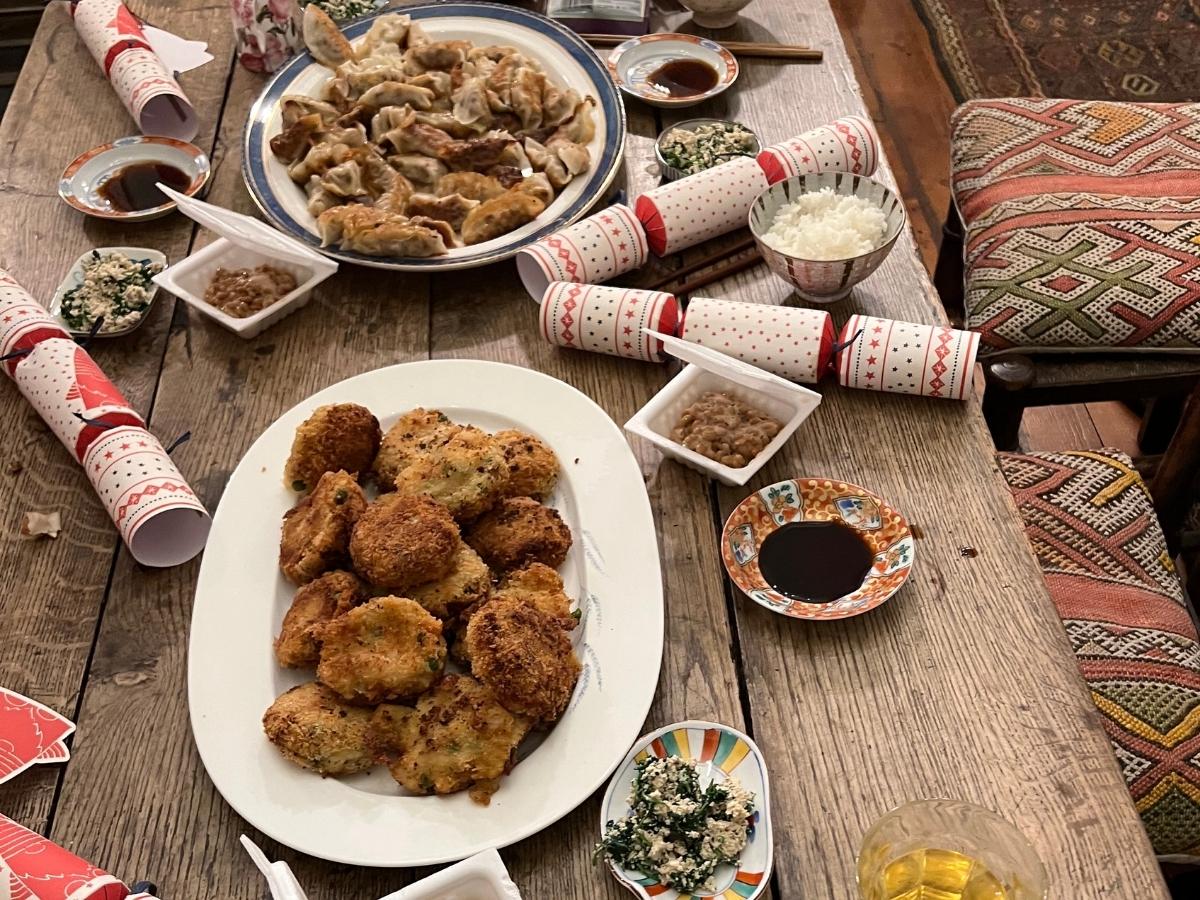 Family Christmas table with plates of Japanese korokke and gyoza, bowls of rice, natto, tofu spinach salad, soy sauce for dipping, and festive crackers.