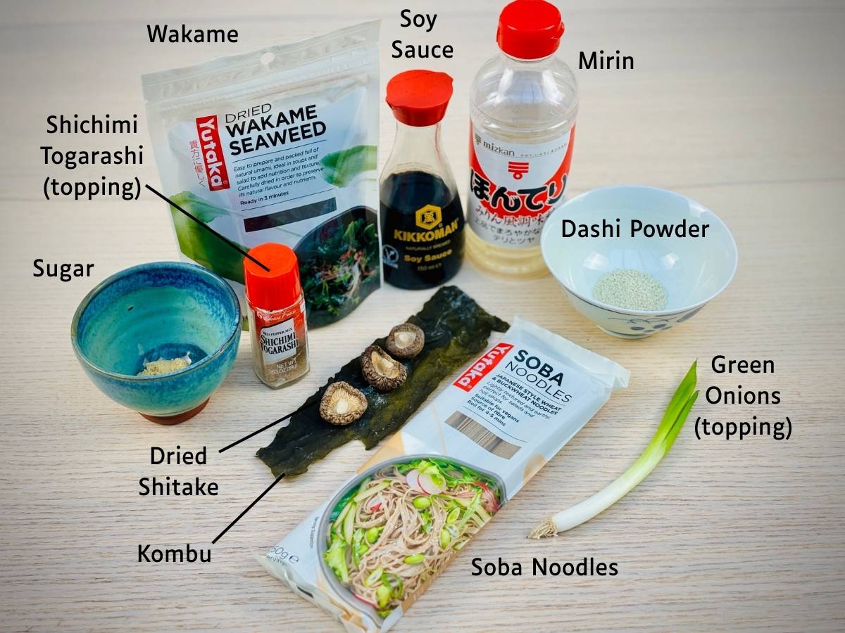 Tempura soba broth ingredients laid out on a worktop and labelled; wakame, soy sauce, mirin, dashi powder, kombu, dried shitake and sugar. Dried soba noodles and toppings, green onions and shichimi togarashi are also on the table.