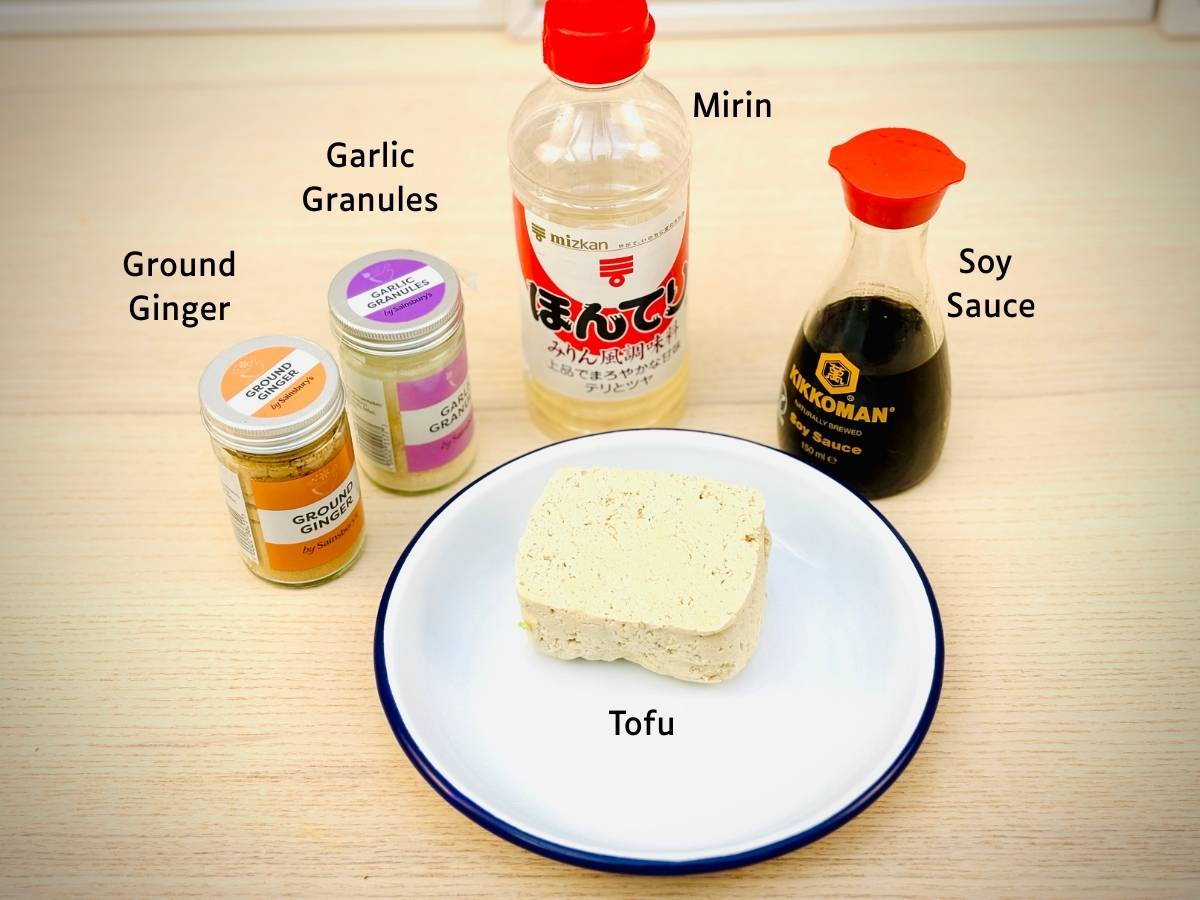 Yaki tofu ingredients laid out on a worktop and labelled; Ground ginger, garlic granules, mirin, soy sauce and tofu.
