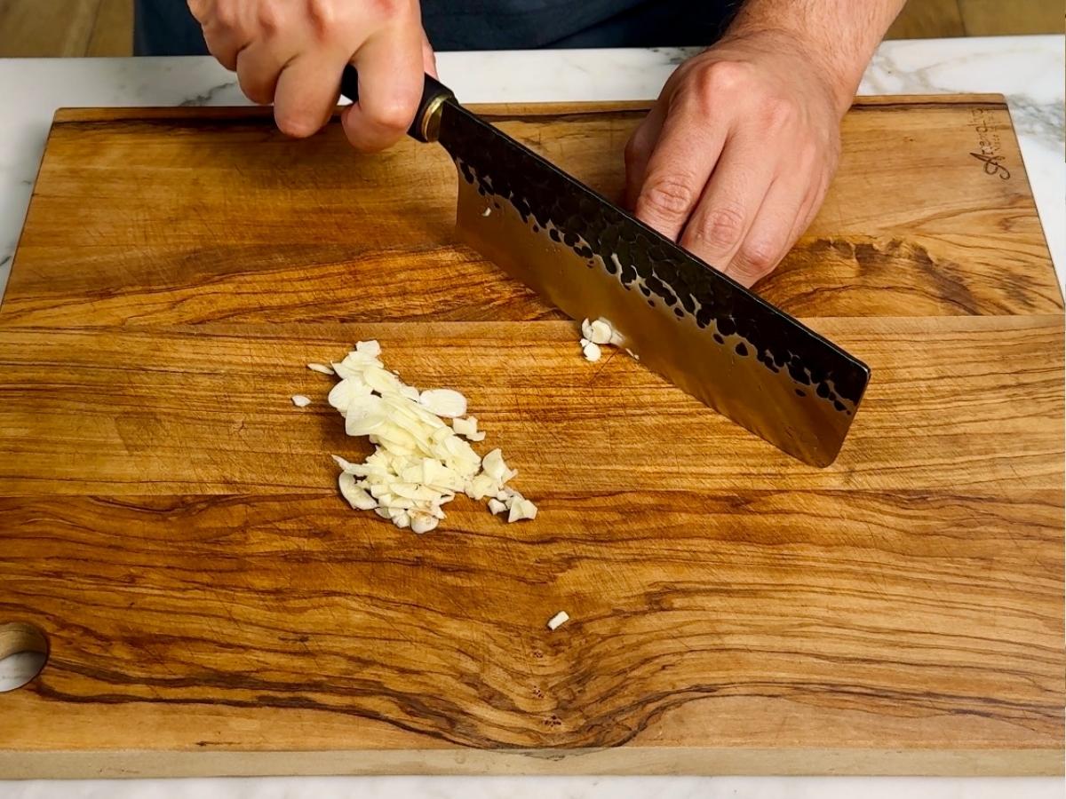 Fresh garlic cloves thinly sliced on a wooden chopping board with a chef’s knife.