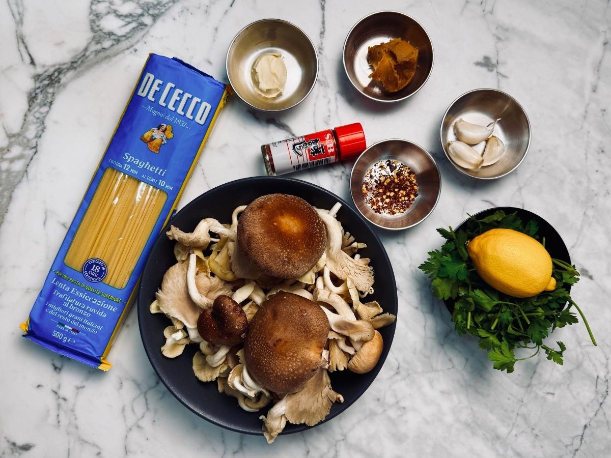 some of the ingredients for miso mushroom pasta pictured: a sealed packet of 'De Cecco' spaghetti, small bottle of shichimi togarashi, a bowl of mixed mushrooms, a small dish of dried chillis,  some miso, some garlic cloves, a bowl of parsely, a knob of butterand a lemon. 