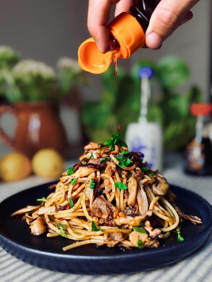 La-yu chillio oil being squeezed out of a bottle by a hand over a finished, plated up portion of miso mushroom spaghetti