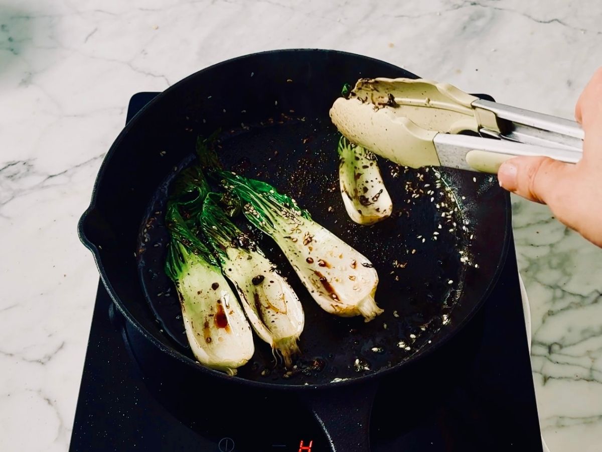 Searing bok choy in soy sauce and mirin while adding sesame seeds to the hot pan.
