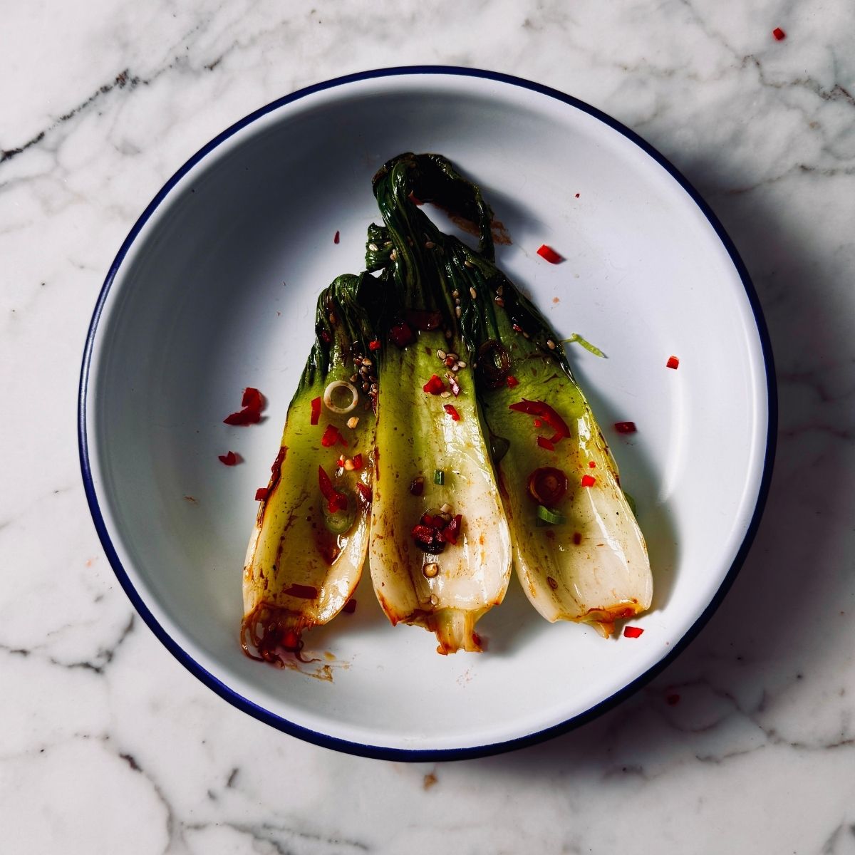 Vegan dish of soy seared bok choy served on a small plate with chili garnish, seasame seeds and green onion toppings