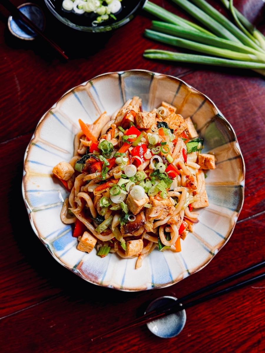 spicy yaki udon next to some spring onion stalks on a wooden dining table