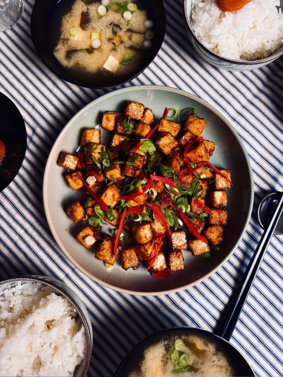 sticky tofu on a round plate garnished with pickled ginger and spring onions next to bowls of rice and miso soup