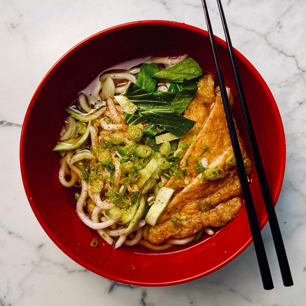 Bowl of vegan udon noodle soup with thick udon noodles in a light vegan dashi broth, topped with inari tofu, pak choi, and spring onions.