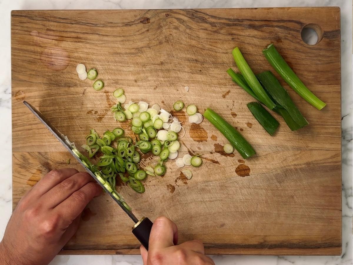 Spring onions being sliced into thin discs on a wooden chopping board