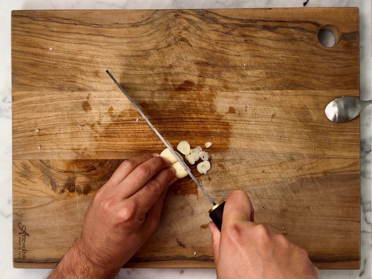 Peeled garlic cloves being sliced into thin discs on a wooden chopping board