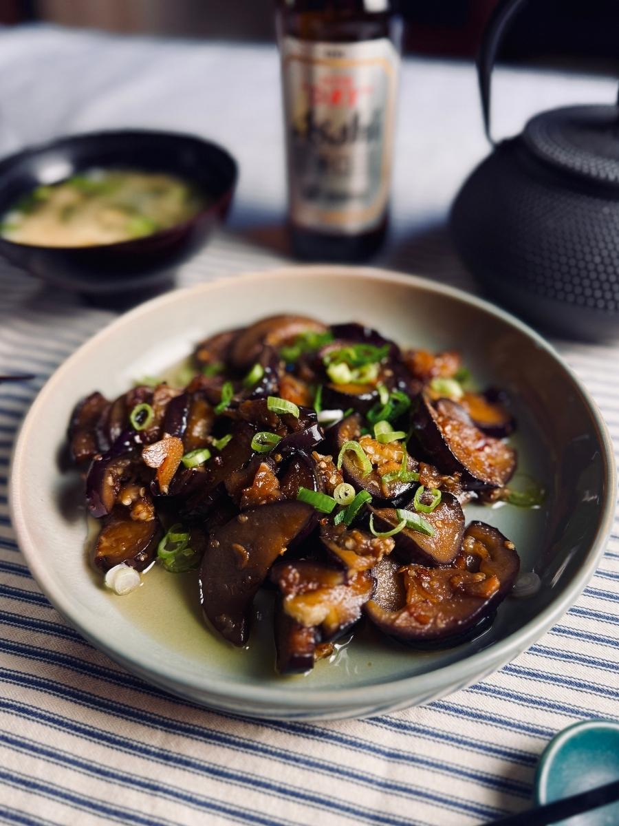 Beer and eggplant stir-fry served in a shallow bowl, garnished with spring onions, with miso soup, Japanese beer, and a teapot in the background.
