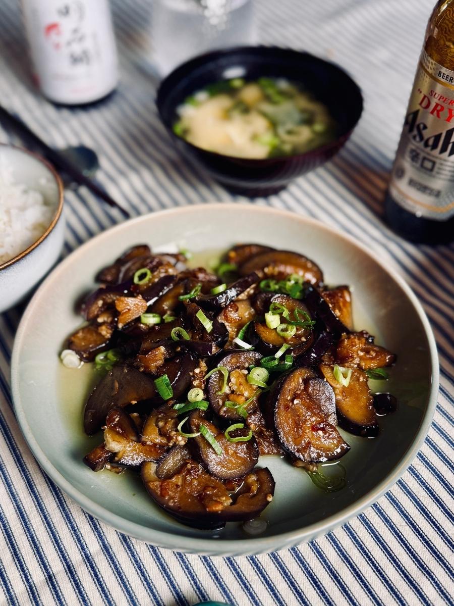 Beer and eggplant stir-fry served in a shallow bowl, garnished with spring onions, with miso soup, Japanese beer, and a teapot in the background.
