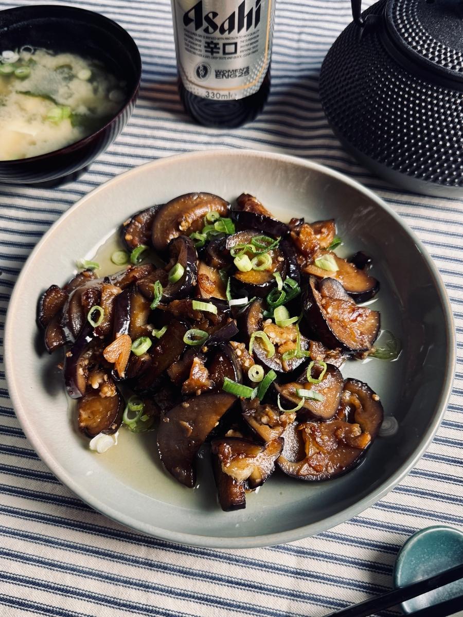 Beer and eggplant stir-fry served in a shallow bowl, garnished with spring onions, with miso soup, Japanese beer, and a teapot in the background.